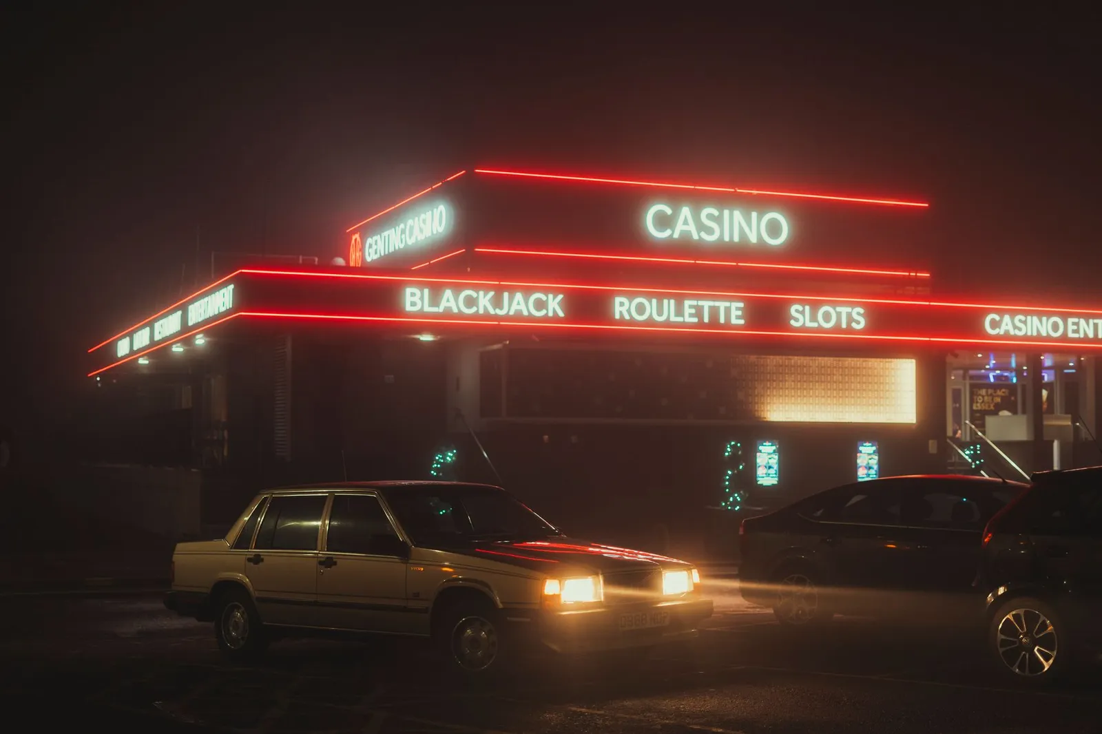 A neon-lit casino stands behind parked cars on a foggy night.